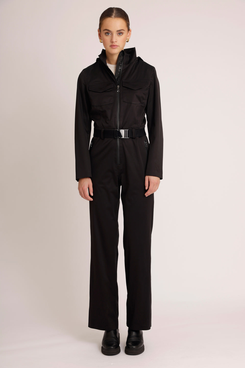 A model wearing a black waterproof stretchable jumpsuit with a hood, paired with black boots and standing against a plain background.