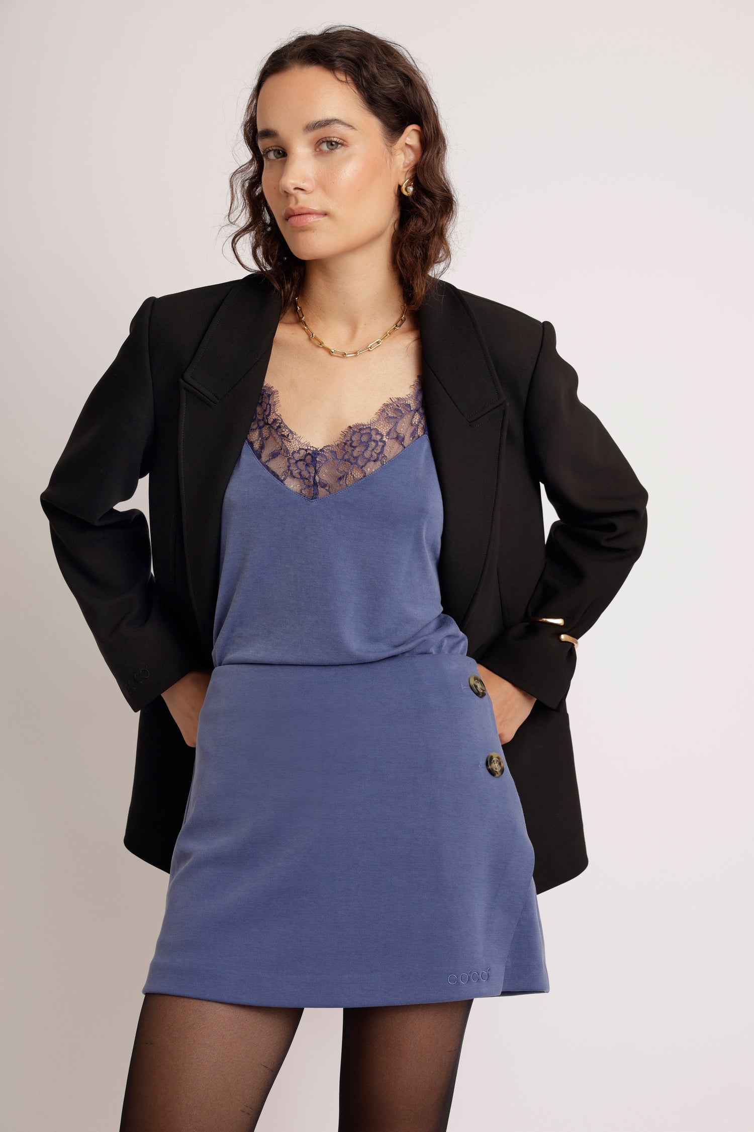 Woman wearing a blue skort with lace detailing camisole and a black blazer on a plain background
