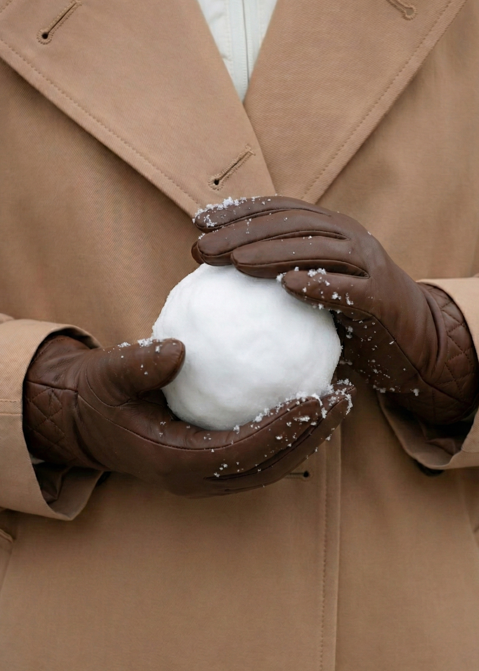 Person wearing a tan coat and brown gloves holding a snowball.