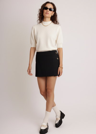 Woman wearing a white sweater, black skort, and sunglasses on a plain background